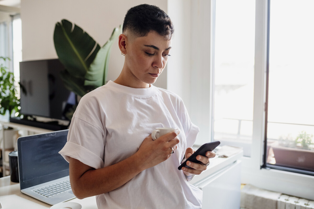 young adult holding a cup of coffee and looking at their smartphone